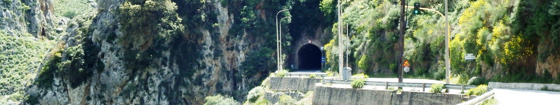 Coastal road leads to a dark tunnel carved in a rocky cliff, with guardrails and green vegetation on both sides.