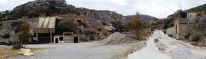 Abandoned quarry with concrete buildings, rubble, and a cracked road at the base of a rocky hillside under a cloudy sky.