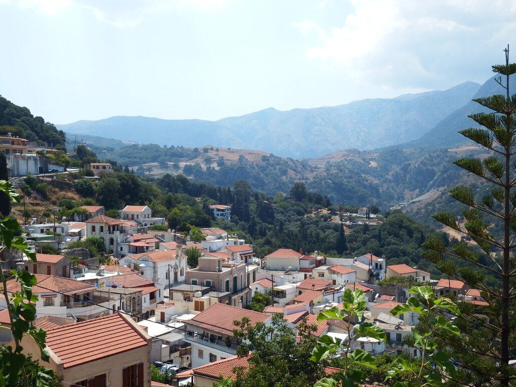 Hillside town with white buildings and red-tiled roofs amid green vegetation and distant mountains under a bright sky.